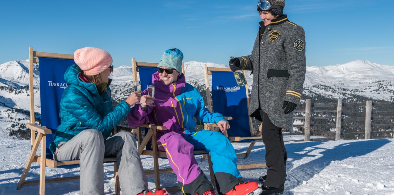 Zwei Skifahrerinnen lassen sich bei Sonnenschein vom Pistenbutler mit Sekt versorgen Pistenbutler-Service